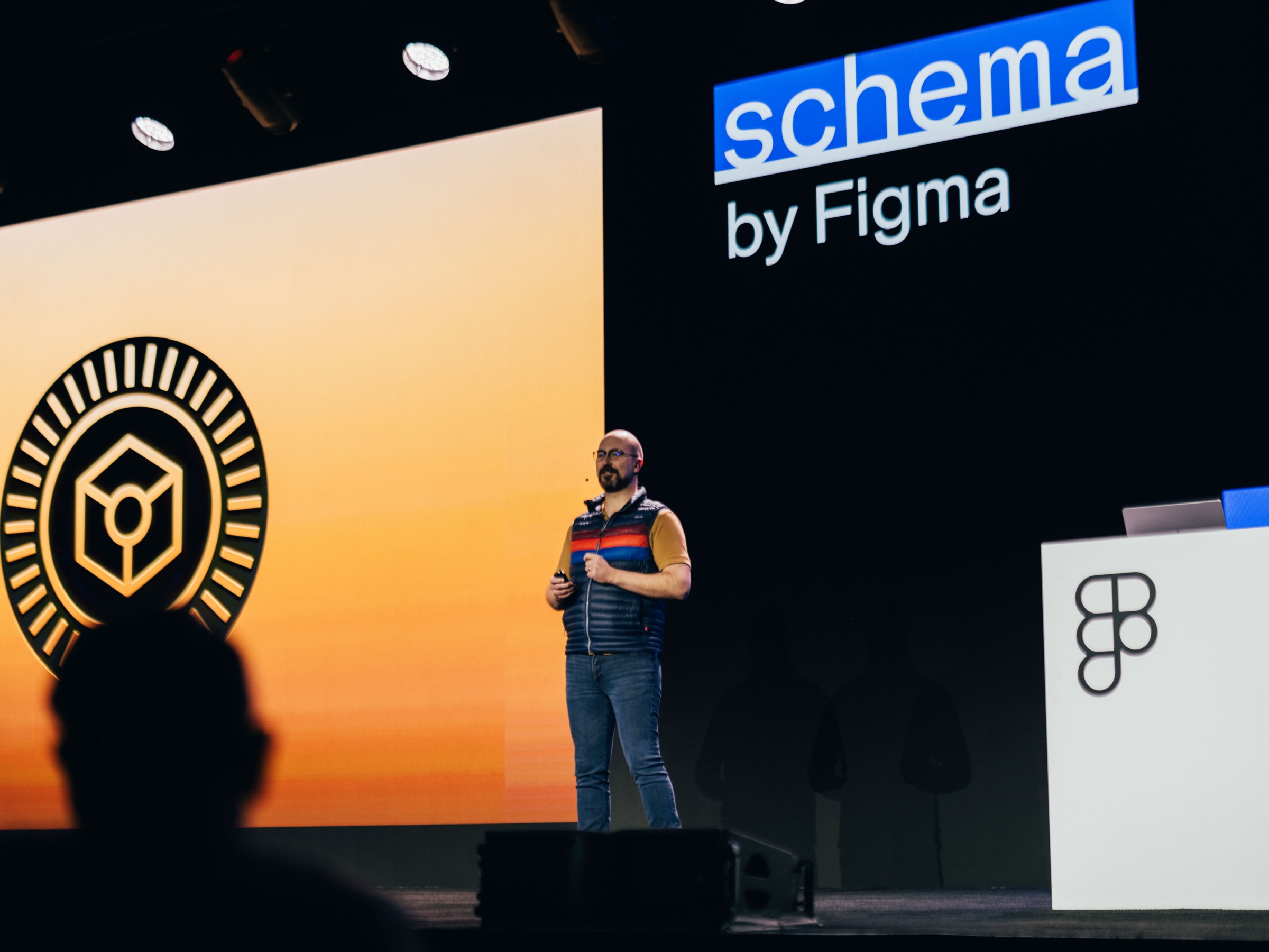 Kaelig speaking on stage at Schema by Figma, in a blue vest, with a warm orange slide behind.